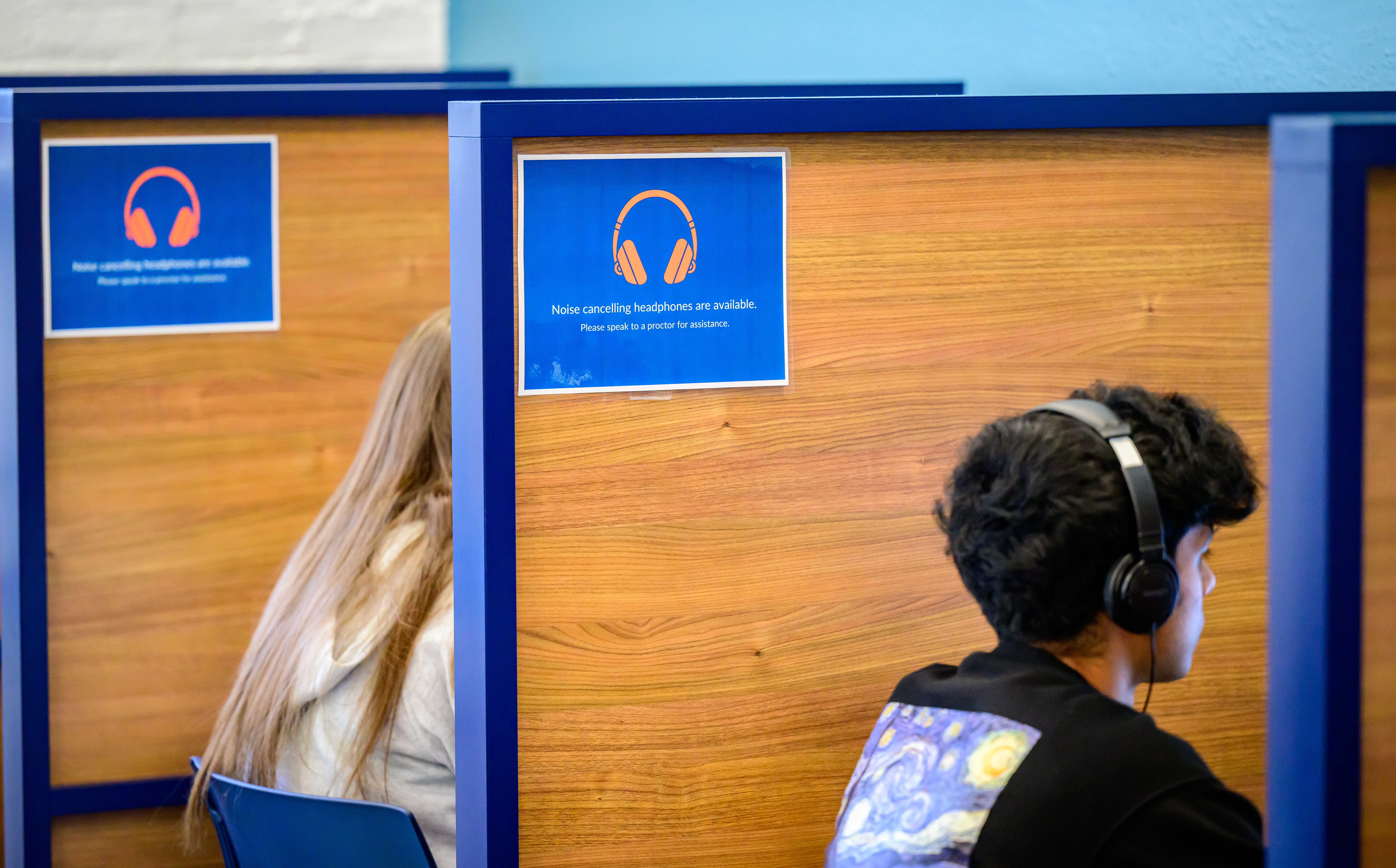 Students sit at computer desks that have wooden divider panels between the. A sign says, &amp;quot;Noise cancelling headphones are available. Please speak to a proctor for assistance.&amp;quot;