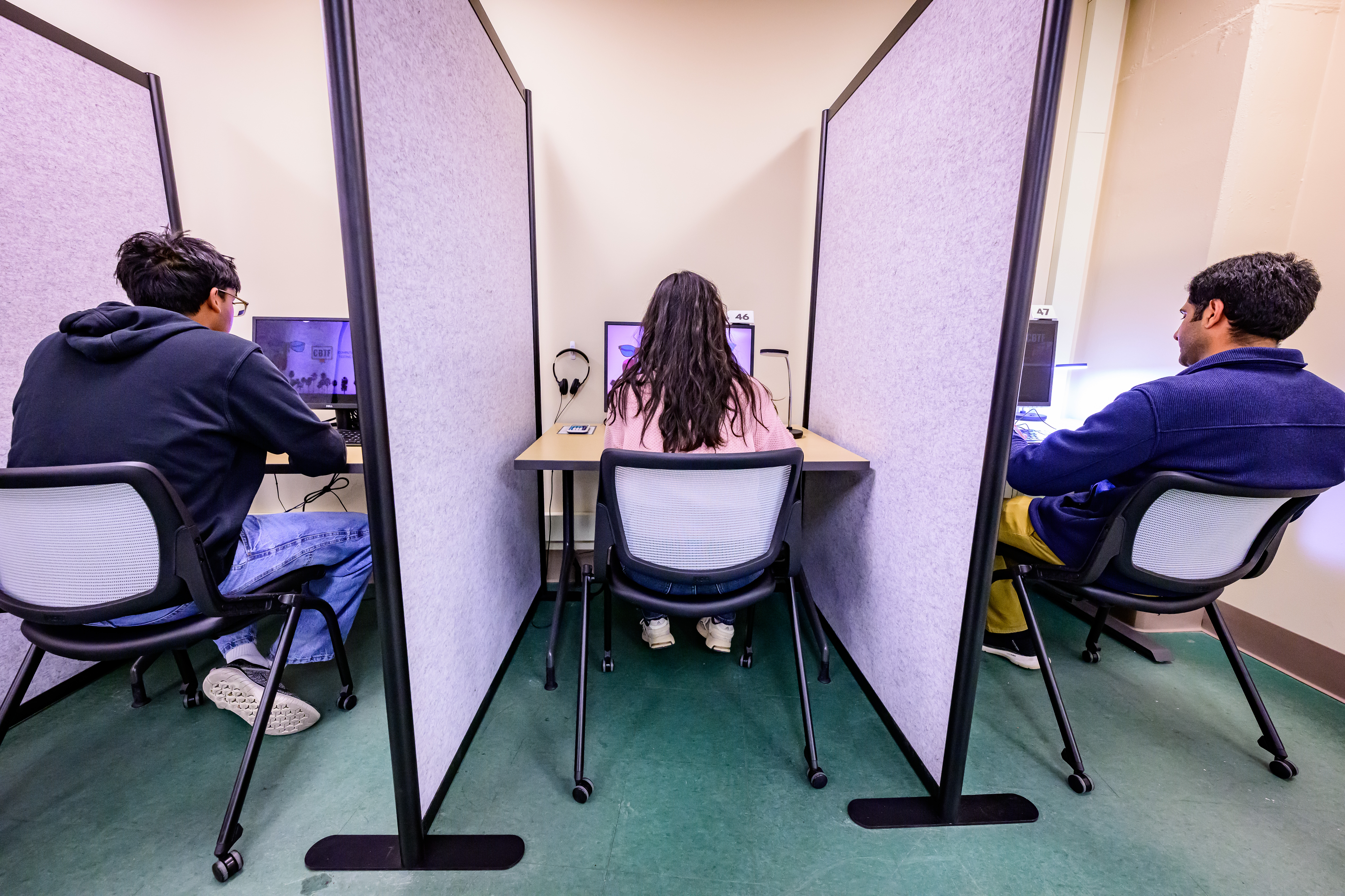 Students sit at computer desks with dividers between them.